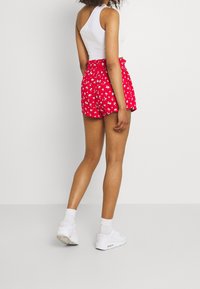 Pantaloni corti rossi a fiori con vita smock, abbinati a una maglietta bianca senza maniche e a sneakers bianche. Il materiale sembra leggero e informale.
