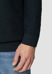 Schwarzer strukturierter Strickpullover mit geripptem Detail am Bündchen, getragen vor einem hellblauen Denim-Hintergrund, mit einer Hand, die an dem Stoff ruht.