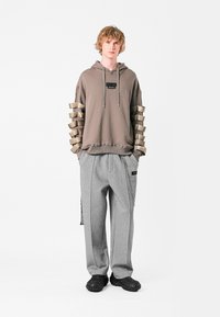 Sudadera gris con detalles de tirantes asimétricos en beige en las mangas, combinada con pantalones grises holgados que presentan cremalleras y acentos de logo, y zapatos negros texturizados.