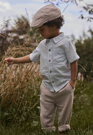 Jeune enfant portant une casquette beige, une chemise bleu clair et un pantalon beige, debout dans un champ herbeux, touchant l'herbe haute d'une main.