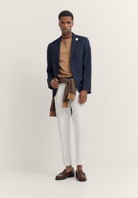 Blazer blu navy su un maglione marrone, pantaloni bianchi e mocassini marroni. Maglione a righe marroni e beige annodato in vita. Sfondo bianco.