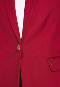 Gros plan d'un blazer rouge avec un bouton décoratif doré et un petit détail en forme de cœur doré sur le rabat de la poche, au-dessus d'un vêtement blanc.