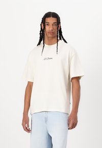 Cream-colored oversized cotton t-shirt with short sleeves featuring black cursive "Le Pratin" text on the chest. Worn with light blue jeans.