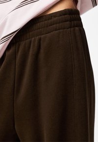 Zbliżenie na ciemno brązowe spodnie dresowe z elastycznym pasem, noszone z włożoną do środka różowo-czarną koszulą w paski.