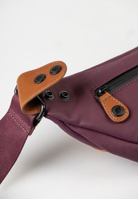 Borsa di stoffa color bordeaux con un accento in pelle marrone, dotata di chiusura a zip e accessori circolari. Inclusa una tracolla regolabile.