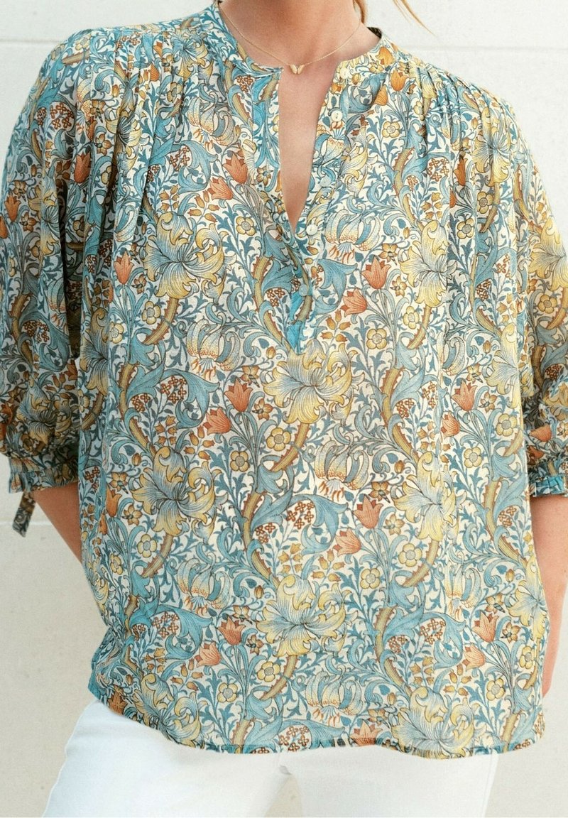 Femme portant un chemisier ample à motif floral avec des fleurs bleues, jaunes et oranges et un pantalon blanc sur un fond neutre.
