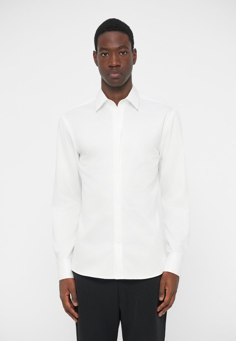 White long-sleeve shirt made of smooth fabric, featuring a pointed collar, button front, and cuffs, worn with dark trousers.