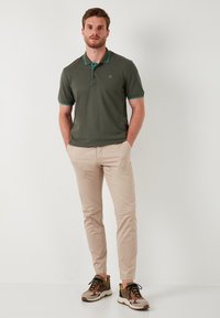 Temnozeleni polo majica s turkiznimi detajli, v kombinaciji s khaki chinos hlačami in supergami v kamuflažnem vzorcu. Preprost dizajn z vsakodnevnim krojem.