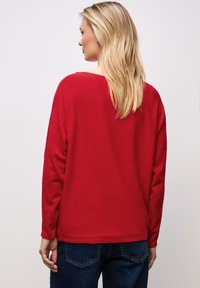 Rotes langärmliges Oberteil mit entspannter Passform, scoopförmigem Rückenausschnitt und weichem Material, kombiniert mit dunkelblauen Jeans.