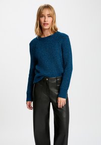 Teal gebreide trui met een textuurpatroon en een wijde halslijn, gecombineerd met zwarte leren high-waisted broeken.