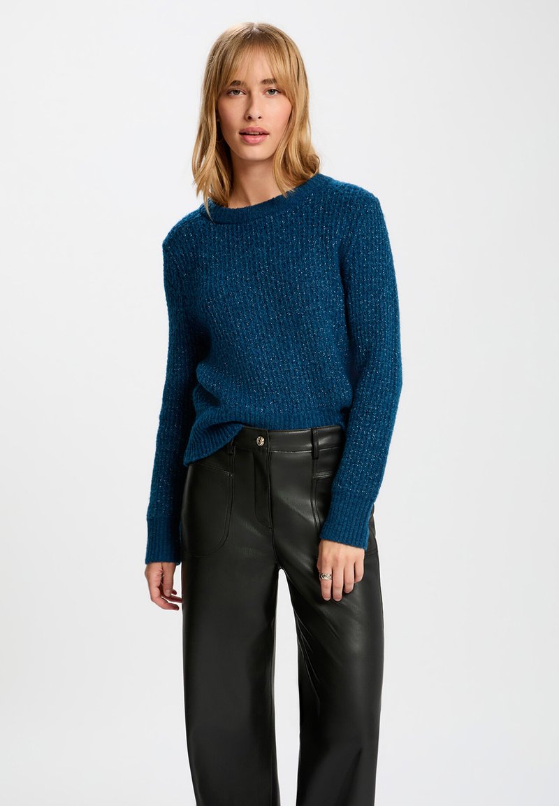 Teal gebreide trui met een textuurpatroon en een wijde halslijn, gecombineerd met zwarte leren high-waisted broeken.