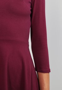 Burgundy mekko, jossa on istuva yläosa ja kolmivartiset hihat; valmistettu sileästä, joustavasta kankaasta; levenevä hame.