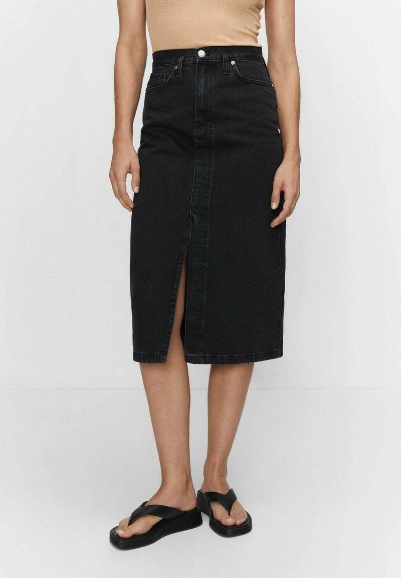 Femme portant une jupe en denim noire longueur genou avec une fente devant, un haut beige sans manches, et des sandales compensées noires, debout sur fond blanc.