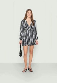 Vestido preto envolvente com padrão de diamante branco, mangas longas e cintura ajustada. Combinado com sapatos de salto preto com tiras e uma pequena mala preta.