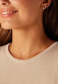 Beige geribbelde trui met een ronde halslijn, gecombineerd met zilveren oorbellen met kleine steentjes en subtiele sprankelende accenten.
