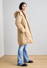 Lys brun polstret parka med hette, med glidelåslommer på sidene og rett snitt. Båret over vide blå jeans og svarte sko.