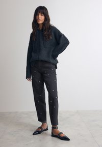 Grauer gestrickter Kapuzenpullover mit Kordelzügen, kombiniert mit schwarzen eng zulaufenden Hosen mit silbernen runden Verzierungen und schwarzen flachen Schuhen.