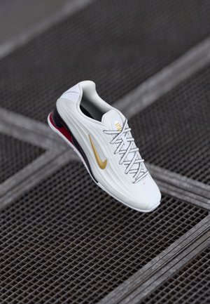 Hvide og guld sneakers med snørebånd svævende over et metalgittergulv i en urban indstilling.