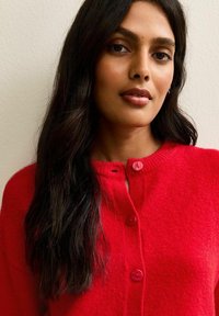 Rode gebreide cardigan met een ronde halslijn, voorzien van twee roze knopen en een zachte textuur. Het kledingstuk heeft een getailleerde pasvorm en geribbelde manchetten.