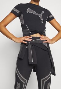 Svart träningslinne med knuten front, med grå Puma-logotyp och geometriska mönster. Svarta leggings med kontrasterande sidodetaljer och knytbart midjeband.