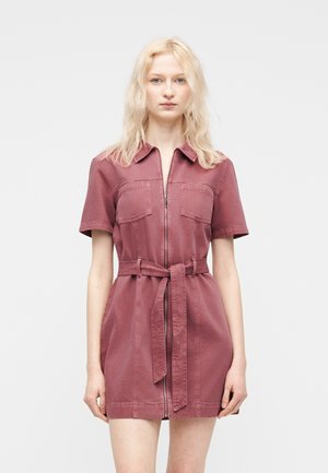 Lyshåret kvinde iført en kortærmet, støvet rosa kjole med lynlås foran, lommer og et bundet bælte i taljen, stående mod en ensfarvet baggrund.