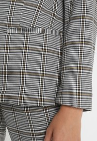 Mustavalkoinen houndstooth-kuvioinen kangas, jossa on ohuita keltaisia raitoja, sileällä tekstuurilla ja löysällä mittasuhteella, näyttäen hiha- ja helmapohjan yksityiskohtia.