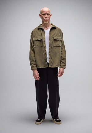 Stojaci plešatý muž oblečený v olivovo zelenej zapínanej bunde, bielej košeli, čiernych nohaviciach so širokými nohavicami a čiernych teniskách s bielymi podrážkami.