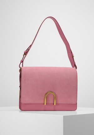 Sac à main en daim rose avec bandoulière réglable et boucle métallique dorée en forme de U sur le rabat avant, présenté sur un socle blanc.