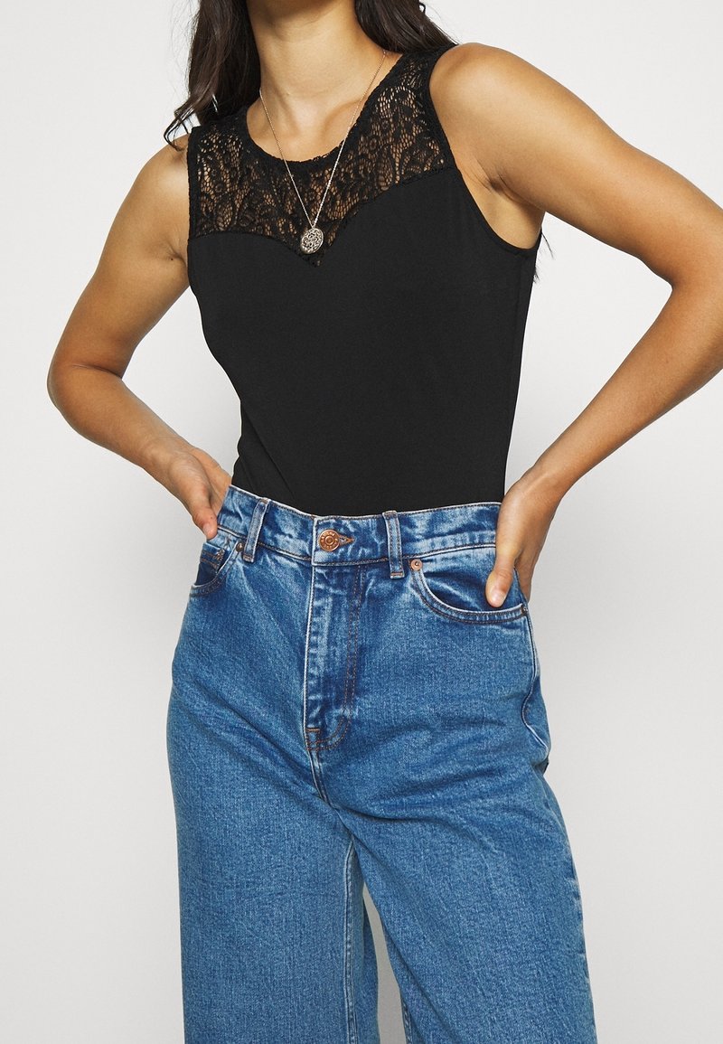 Femme portant un débardeur noir avec un détail en dentelle et un jean bleu taille haute, les mains posées sur les hanches.