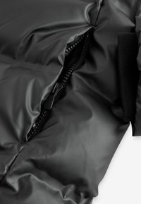 Chaqueta acolchada negra con una textura brillante y suave. Cuenta con una cremallera corta y acentos elásticos a lo largo de los bordes.