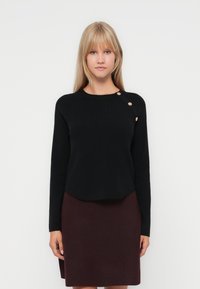 Pull noir à manches longues et encolure ronde, orné de trois boutons dorés sur l'épaule gauche. Associé à une jupe bordeaux.