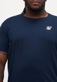 Tricou din bumbac de culoare bleumarin, cu mâneci scurte, decolteu rotund și logo brodat pe pieptul stâng. Textură netedă și design simplu.
