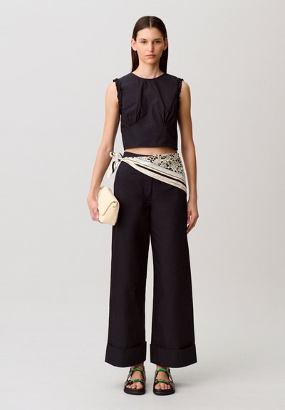Top nero corto con maniche arricciate, pantaloni neri larghi, sciarpa bianca con motivo in vita e una piccola borsa crema; sandali con dettagli verdi.