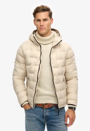 Pufferjack in lichtbeige met een gestructureerde afwerking, capuchon en zwarte rits. Draagt men over een crèmekleurige coltrui en blauwe spijkerbroek met een bruine riem.
