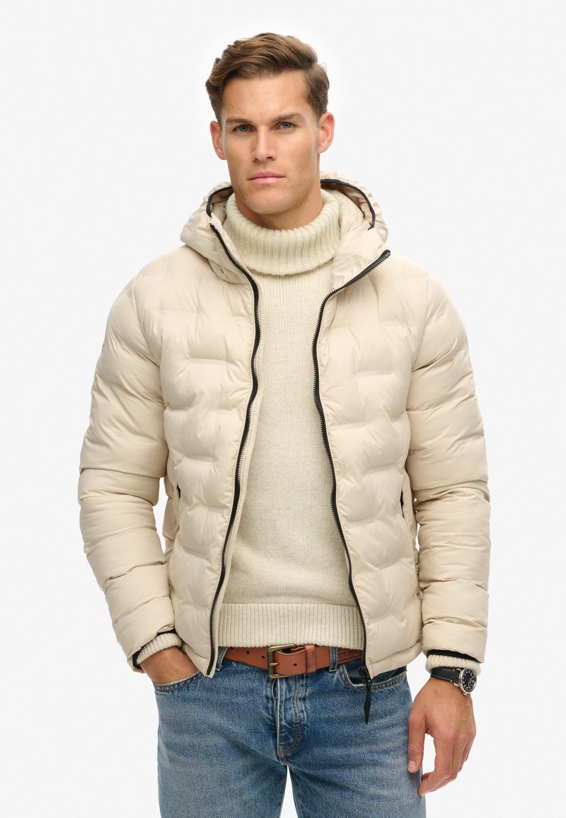 Pufferjakke i lyse beige med en tekstureret overflade, hætte og sort lynlås. Båret over en cremet turtleneck og blå denimjeans med et brunt bælte.