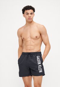 Shorts de bain noirs avec une taille à cordon, arborant un logo blanc "GALLIAN KLEIN" verticalement sur le côté gauche. Texture de tissu lisse.