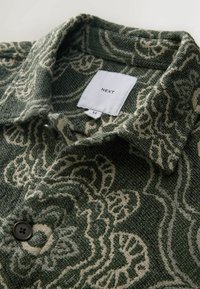 Green patterned collared shirt featuring intricate cream floral designs, dark buttons, and a label indicating size M. Textured fabric.