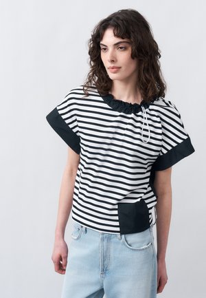 Γυναίκα με σγουρά σκούρα μαλλιά φοράει πουκάμισο με ασπρόμαυρες ρίγες και μαύρα μανίκια καθώς και ανοιχτόχρωμο μπλε τζιν, στέκεται μπροστά σε ένα μονόχρωμο φόντο.