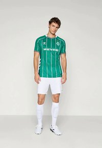 Zaļš futbola krekls ar vertikālām baltām svītrām, ar logotipu un tekstu. Apvienots ar baltām šortiem un zeķēm. Vieglas, sportiskas piegriezums.