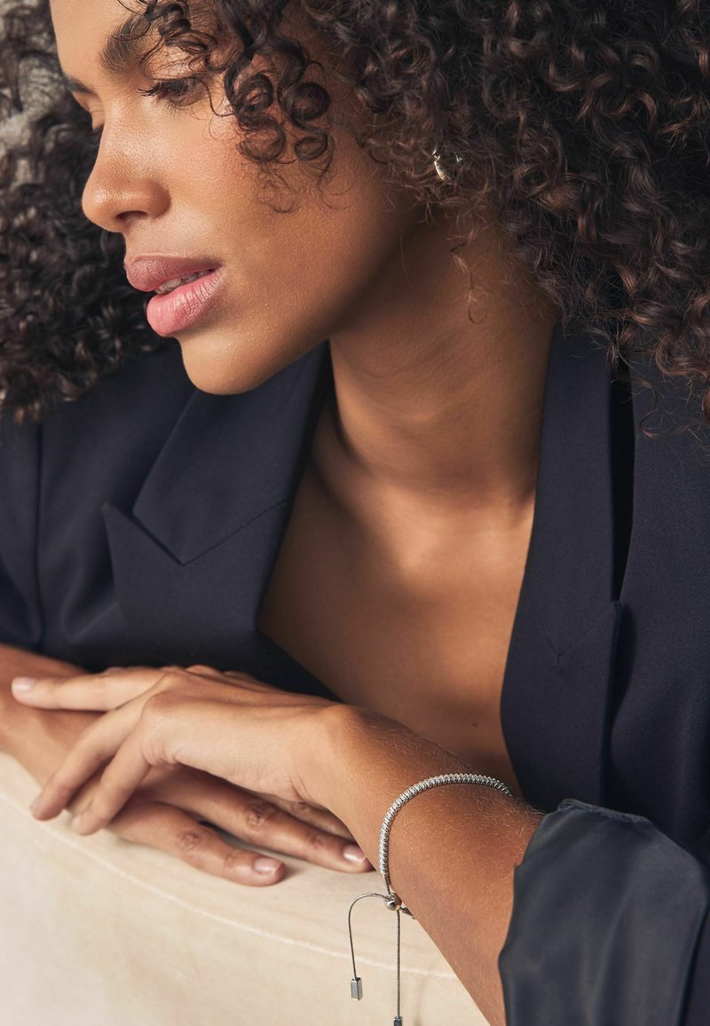Gros plan sur le visage d'une femme portant un blazer noir, avec des cheveux bouclés et un bracelet argenté à son poignet, reposant sur une surface de couleur claire.