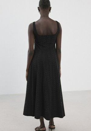 Mujer de pie de espaldas a la cámara, vistiendo un vestido midi negro sin mangas con ojales y sandalias negras, sobre un fondo blanco liso.