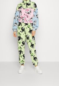 Fleece-Pullover mit rosa und blauen Abschnitten, mit Cartoon-Designs. Passende grüne Hose mit schwarz-weißen Mustern und Herzen. Weiße Sneaker.