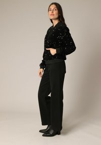 Veste bomber en sequins noirs avec des poignets côtelés, associée à un pantalon noir taille haute et des bottines noires à talons aiguilles. Texture lisse et design ajusté.