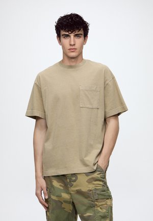 Jeune homme aux cheveux bouclés foncés portant un t-shirt beige avec une poche poitrine et un pantalon cargo camouflage vert, debout avec une main dans la poche.