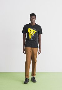 T-shirt preta com o texto amarelo "JUST DO IT", combinada com calças castanhas e sapatos pretos, em pé sobre uma superfície verde em um fundo neutro.