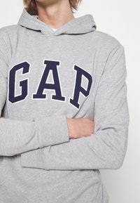 Persona che indossa una felpa grigia chiara con lettere "GAP" blu navy, braccia incrociate, su uno sfondo bianco uniforme.