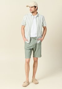 Camisa de manga corta a rayas en verde claro y blanco, combinada con pantalones cortos verde claro y zapatos slip-on beige. Una gorra neutra completa el conjunto.
