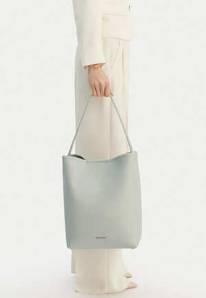 Osoba v světle béžovém obleku drží světle šedou koženou kabelku za jediný ramenní popruh, stojí na jednobarevném bílém pozadí.