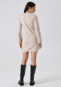 Vestito beige lavorato a maglia con maniche lunghe, scollatura rotonda, bottoni posteriori e una silhouette aderente, abbinato a stivali neri fino al ginocchio.