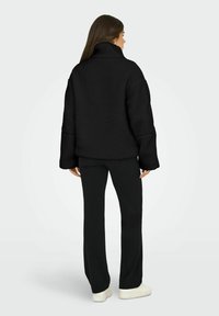Juoda pūkinė striukė su aukštu apykakle, per dideliu kirpimu, tekstūruota paviršiumi ir plačiomis rankovėmis, derinama su juodomis kelnėmis ir šviesiais sportbačiais.